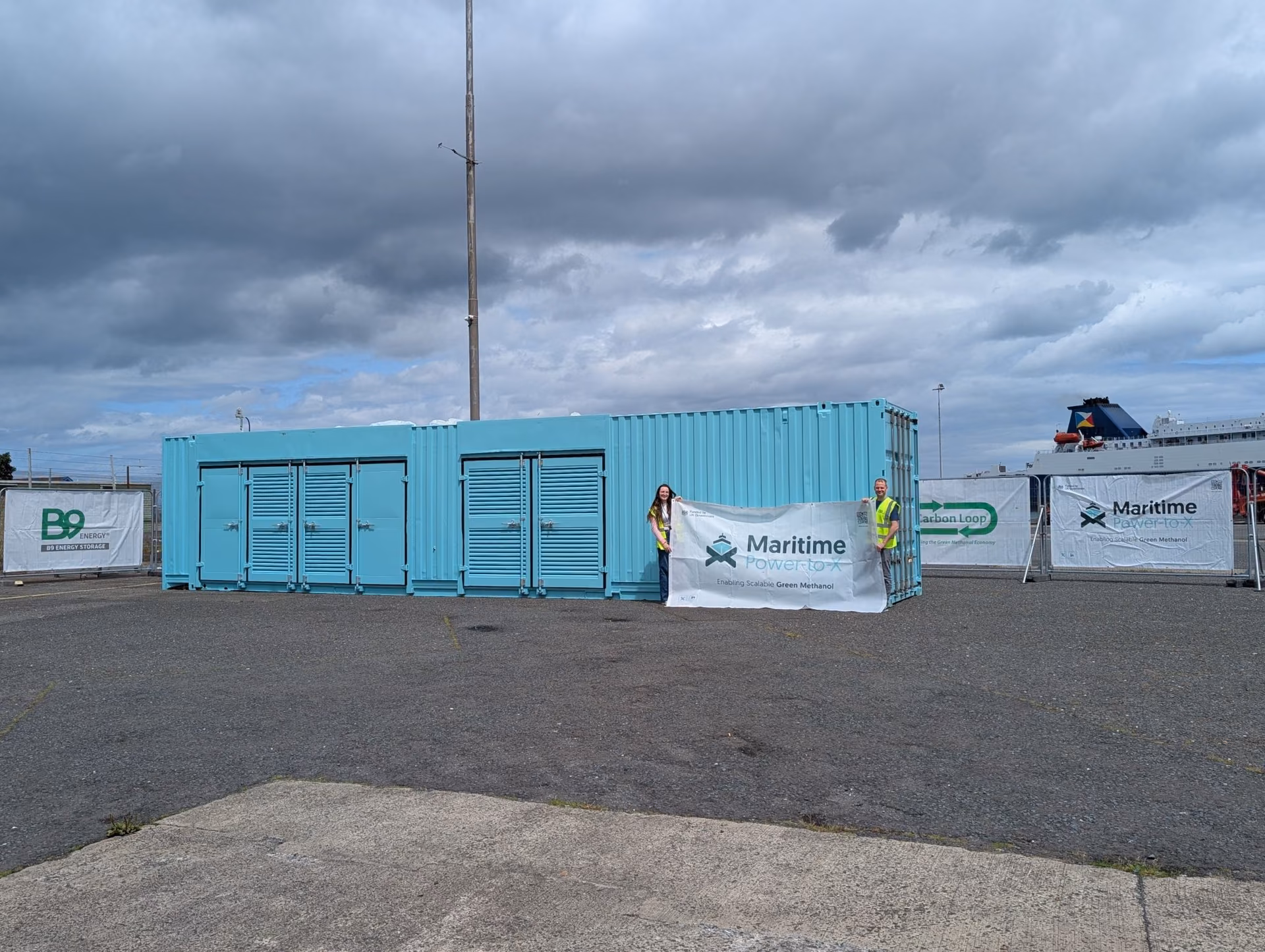 A blue container unit at Larne Harbour with Maritime Power-to-X banners and two people holding a project banner